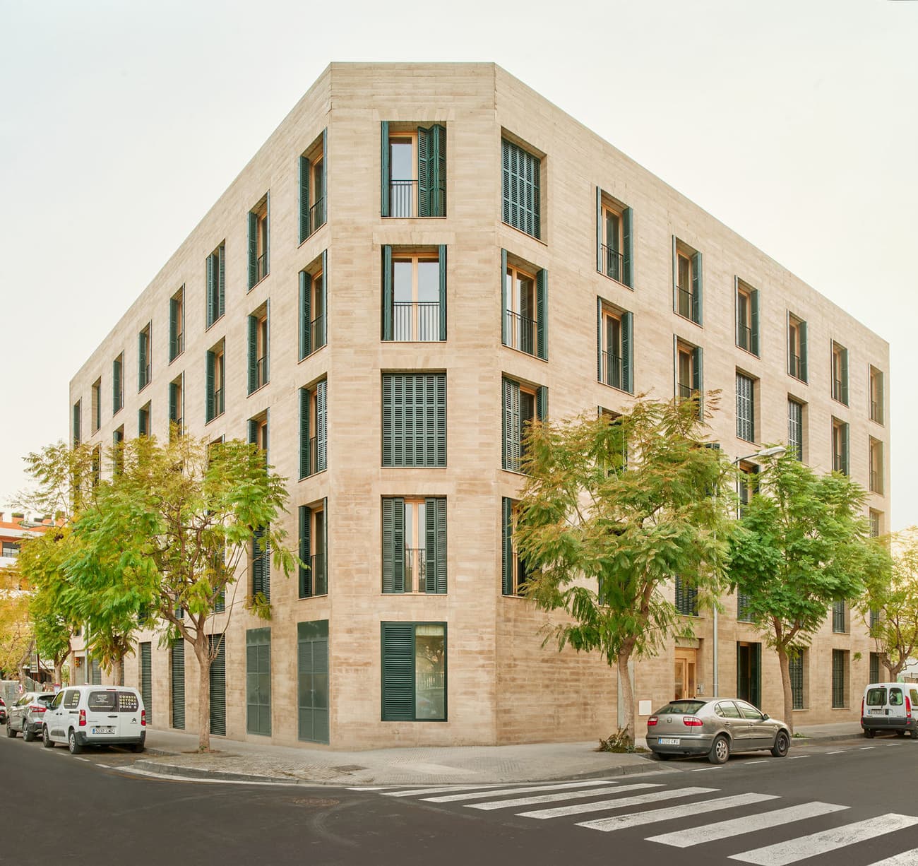 Architectural project - vertical windows, urban greenery, stone facade, apartment block, beige facade, multi-story building, stone cladding, corner building, grid windows, urban residential, eco concrete, natural shading, green urban design, street trees, pedestrian friendly, light stone, green windows, window grid, passive design, sustainable design, warm tones, natural tones, urban landscape, contemporary style, sustainable architecture, neutral tones, urban environment, city building, urban planning, beige, eco friendly, minimalist design, modern facade, clean lines, glass windows, residential building, textured facade, contemporary facade, passive solar, modern style, geometric facade, urban architecture, trees, modern urban, square windows, city dwelling, urban living, street view, urban housing