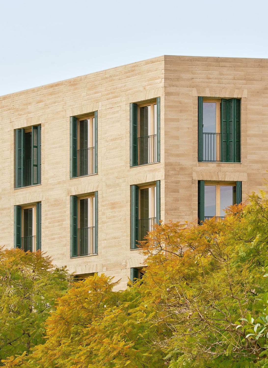 Architectural project - green shutters, modern building, urban residence, stone exterior, beige stone, natural stone, vertical elements, rectangular shape, subtle pattern, multi-story building, green accents, passive shading, refined style, window symmetry, facade depth, eco integration, minimal design, cityscape, sustainable design, warm tones, contemporary architecture, green, beige, daylight optimization, natural textures, minimalist vibe, neutral palette, eco-friendly materials, clean lines, large windows, stone, greenery, nature integration, textured facade, sleek design, urban nature, architectural elegance, facade rhythm, residential block, urban home, urban facade, city dwelling, urban living