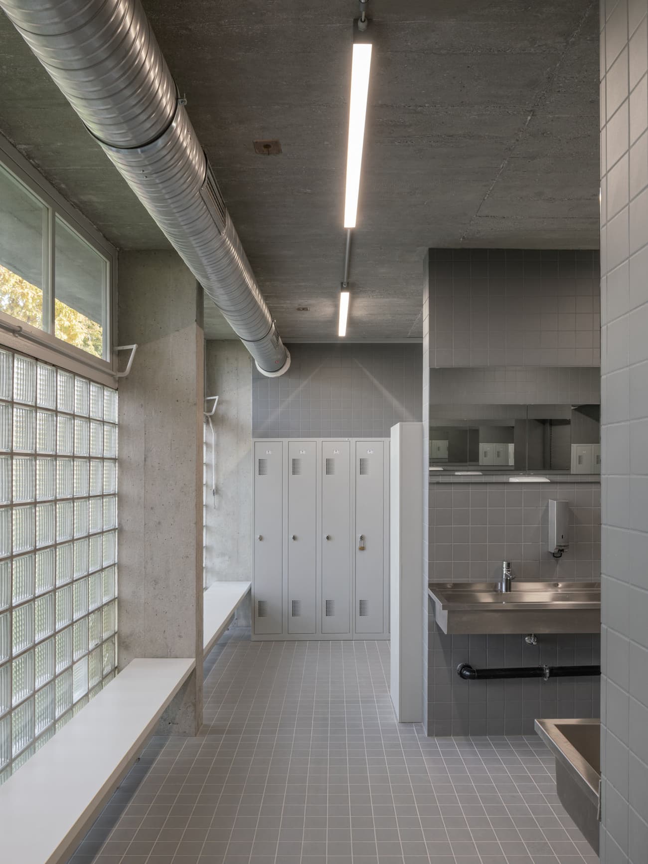 Architectural project - silver, industrial interior, exposed ductwork, commercial space, urban aesthetic, smooth textures, concrete ceiling, concrete walls, steel accents, reflective surfaces, glass bricks, grid pattern, linear lighting, minimalist bathroom, modern fixtures, utilitarian space, public locker room, tiled walls, gray tiles, grey flooring, stainless steel sink, industrial sink, fluorescent lighting, ample storage, locker storage, functional restroom, spatial efficiency, steel, natural light, grey, neutral tones, industrial vibe, concrete, functional design, neutral palette, durable materials, clean lines, light filled, modern architecture, robust design, raw finish, sleek design, airy atmosphere, contemporary space, muted tones, urban style, practical layout