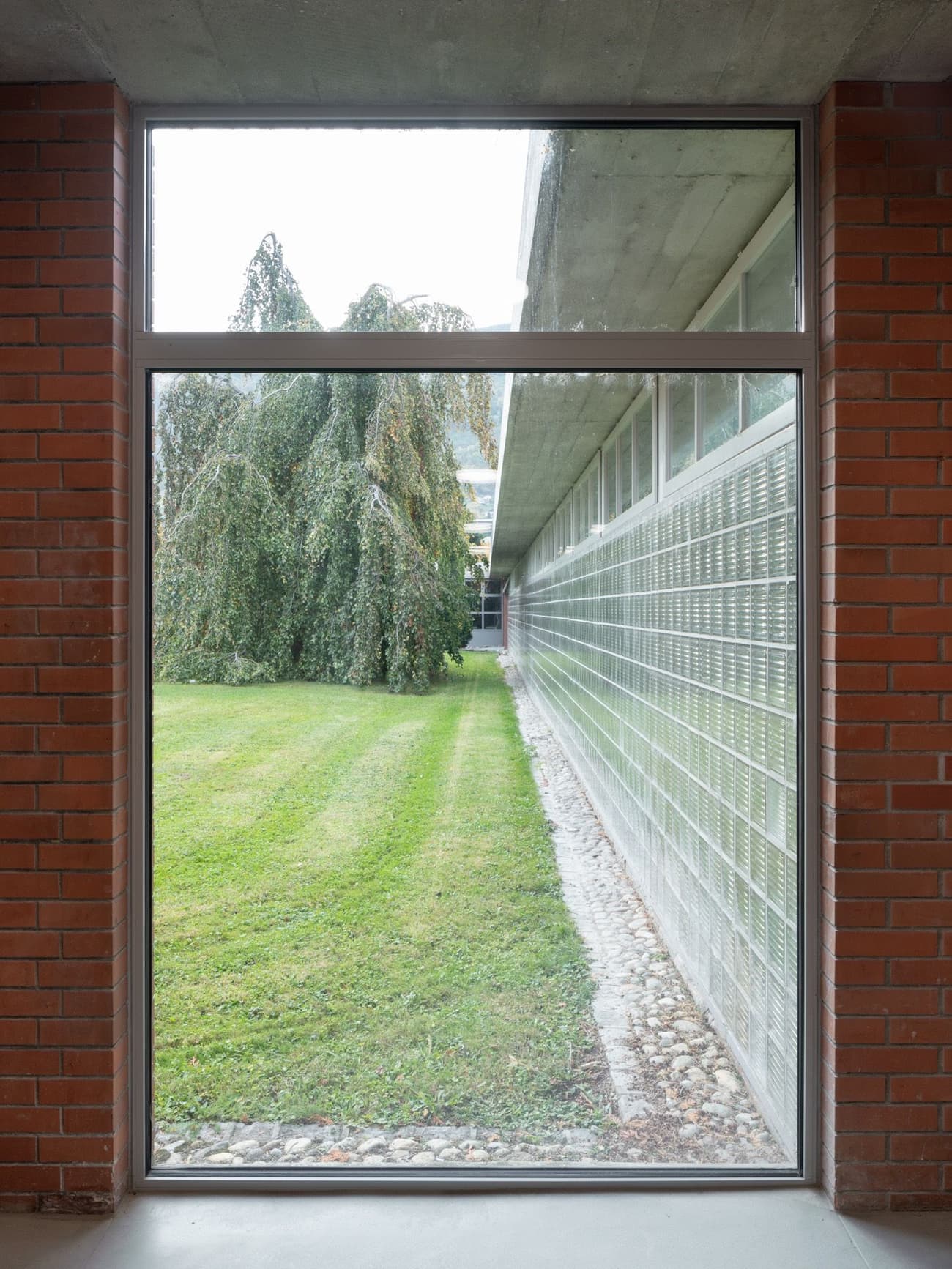 Architectural project - exterior facade, brick wall, landscape, horizontal windows, concrete ceiling, transitional space, glass blocks, lawn, structured space, connection, minimal design, minimalist, architectural detail, industrial design, natural light, sustainable design, glass, warm tones, eco design, grey, green, neutral tones, daylight, industrial vibe, brown, concrete, building facade, industrial style, contemporary, simple design, modern, clean lines, indoor outdoor, large window, glass window, outdoor view, textured wall, framed view, brick, outdoor connection, red brick, garden view, open view, green space, urban style