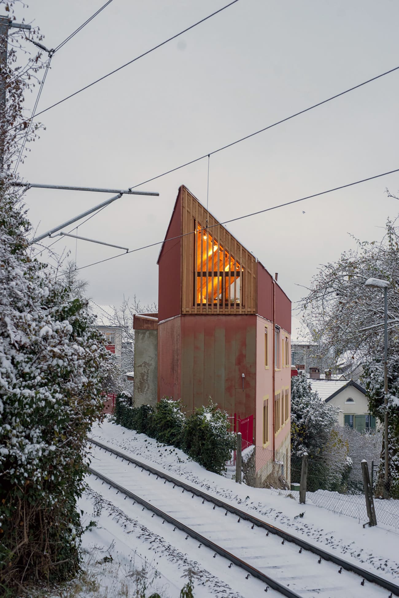 Architectural project - small house, warm glow, colorful exterior, red facade, city home, timber slats, suburban setting, urban house, asymmetrical design, natural materials, wood, timber, natural light, warm tones, eco design, contemporary style, evening light, beige, concrete, modern design, rustic charm, low carbon, sustainable home, passive house, modern architecture, energy efficient, plaster, sloped roof, pitched roof, textured walls, wooden facade, residential building, compact living, urban architecture, vertical cladding, architectural contrast, geometric form, urban retreat, red