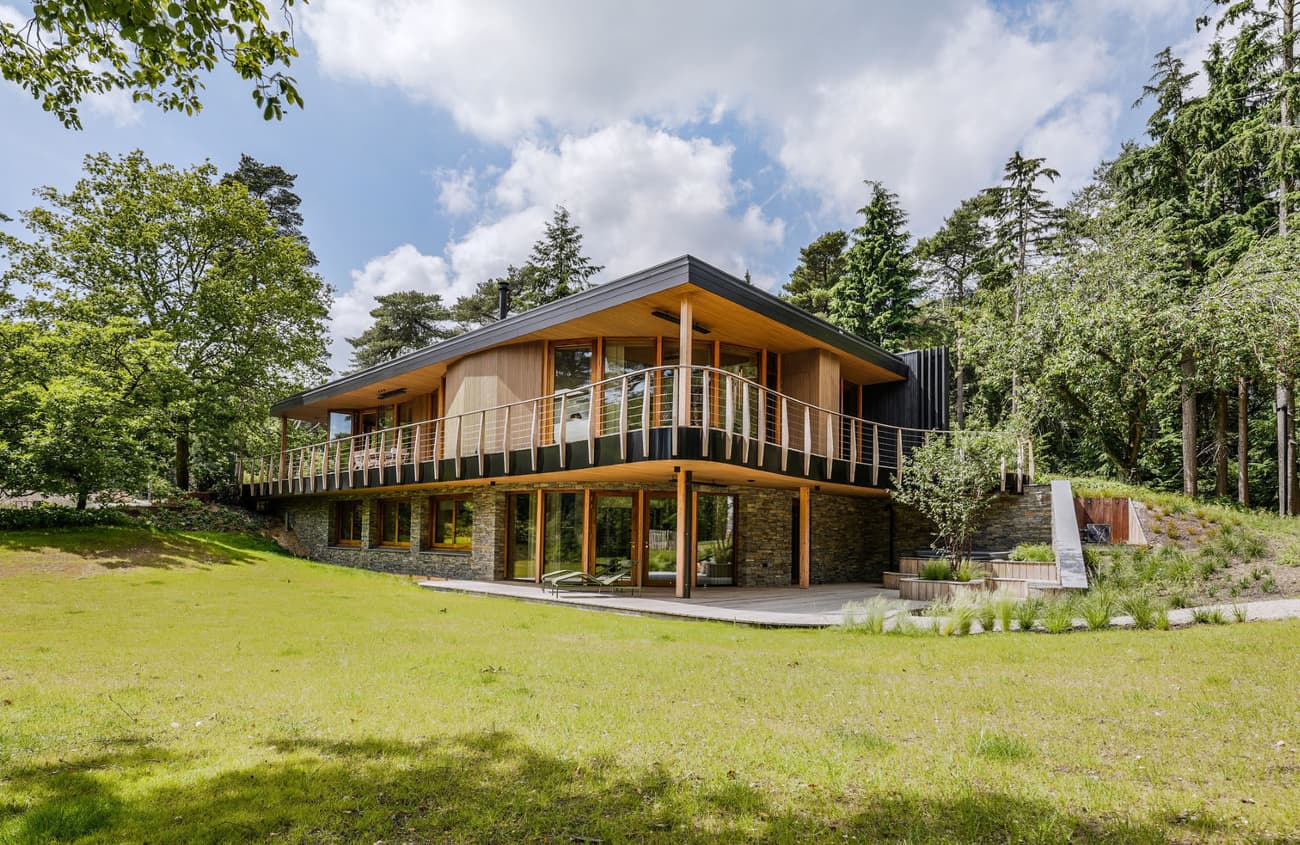 Architectural project - garden patio, stone accents, seamless exterior, hillside home, wraparound balcony, cantilevered design, panoramic windows, daylight home, residential dwelling, idyllic home, natural materials, wood, timber, rustic touch, glass, warm tones, natural tones, eco design, contemporary style, green, beige, timber facade, light wood, eco friendly, wood exterior, natural setting, honey wood, glass doors, tranquil setting, biophilic design, large windows, energy efficient, stone base, stone, sloped roof, forest retreat, forest view, peaceful retreat, landscape design, residential architecture, nature integration, outdoor deck, passive solar, open view, nature blend, green roof, sustainable living, modern cabin, modern retreat, modern house
