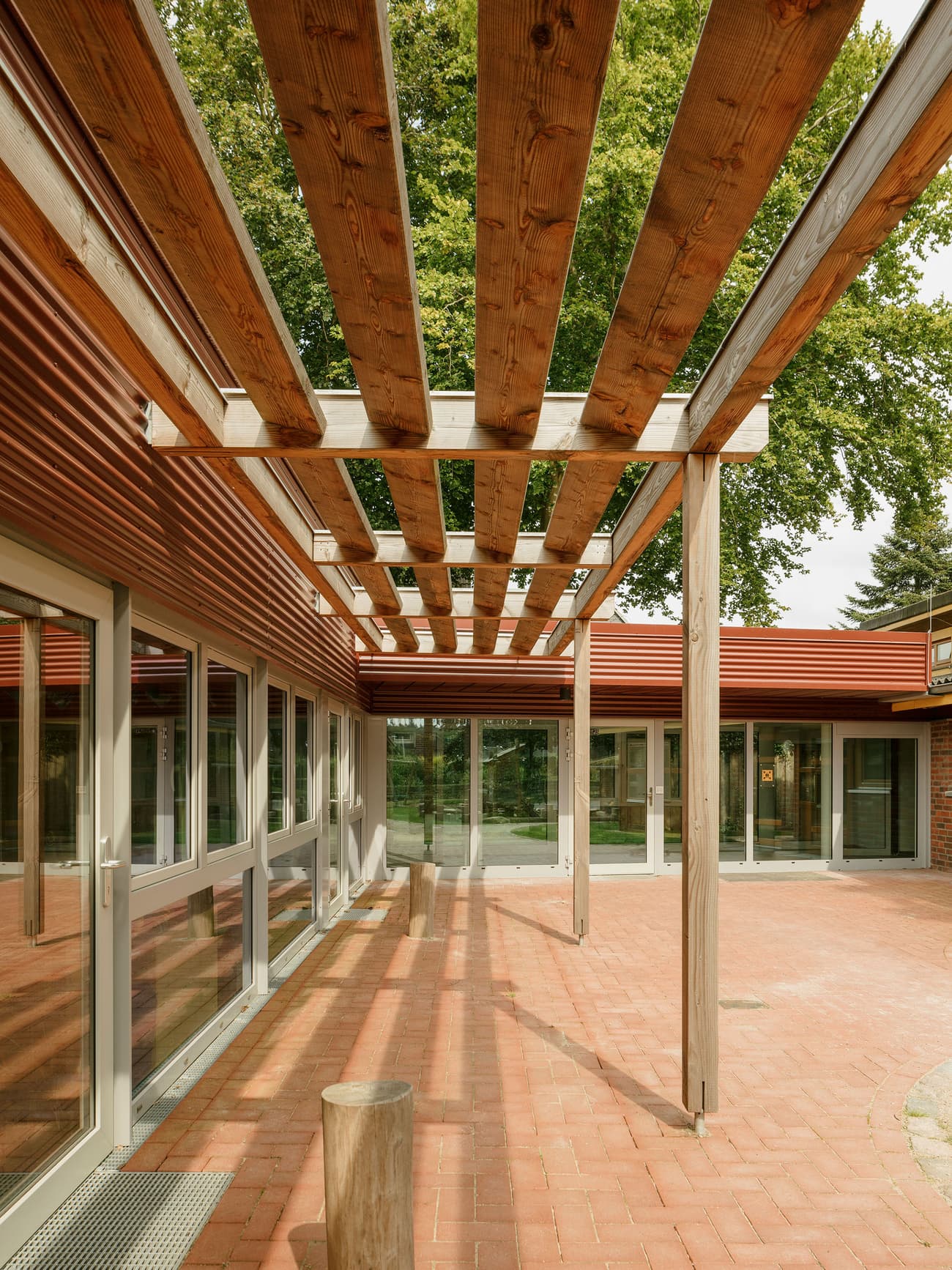 Architectural project - linear design, pergola, brick flooring, communal area, tree shade, natural materials, eco-friendly design, wood, timber, outdoor space, natural light, outdoor living, passive design, glass, warm tones, sustainable construction, community space, contemporary style, neutral tones, daylight, beige, brown, wooden beams, simple design, timber beams, minimalist design, social space, gathering area, glass doors, adaptive reuse, biophilic design, clean lines, modern architecture, inviting space, warm atmosphere, terracotta, brick, greenery, exterior view, tree integration, open air, sliding doors, architectural harmony, courtyard, terracotta floor, red