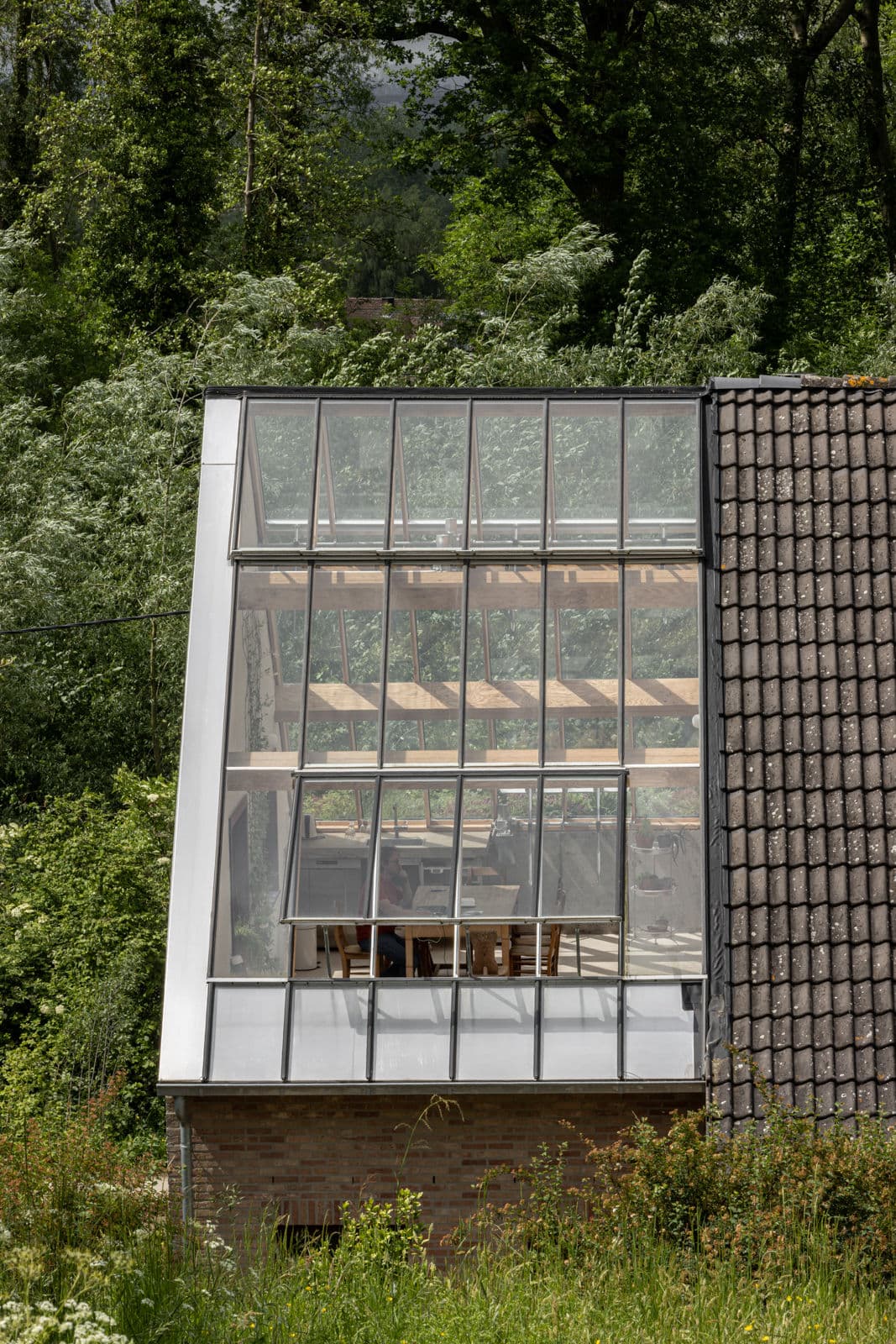 Architectural project - brickwork, transparent facade, grey tiles, roof tiles, brick foundation, window grids, framed glass, daylight design, lush setting, passive solar gain, harmonious blend, textured materials, atmospheric, forest environment, cream tones, terracotta tiles, functional aesthetic, minimal design, natural materials, architectural detail, natural light, warm tones, eco design, sustainable architecture, outdoor setting, exterior design, rural architecture, rustic modern, contemporary design, eco-friendly materials, modern rustic, biophilic design, scenic view, landscape integration, clean lines, light filled, indoor outdoor, modern architecture, countryside home, wood accents, earthy tones, pitched roof, nature-inspired, triangular roof, nature connection, brick base, garden view, cozy design, timber framing, seamless integration, green surroundings, simple lines, glass facade, energy efficiency, glass wall, clay tiles