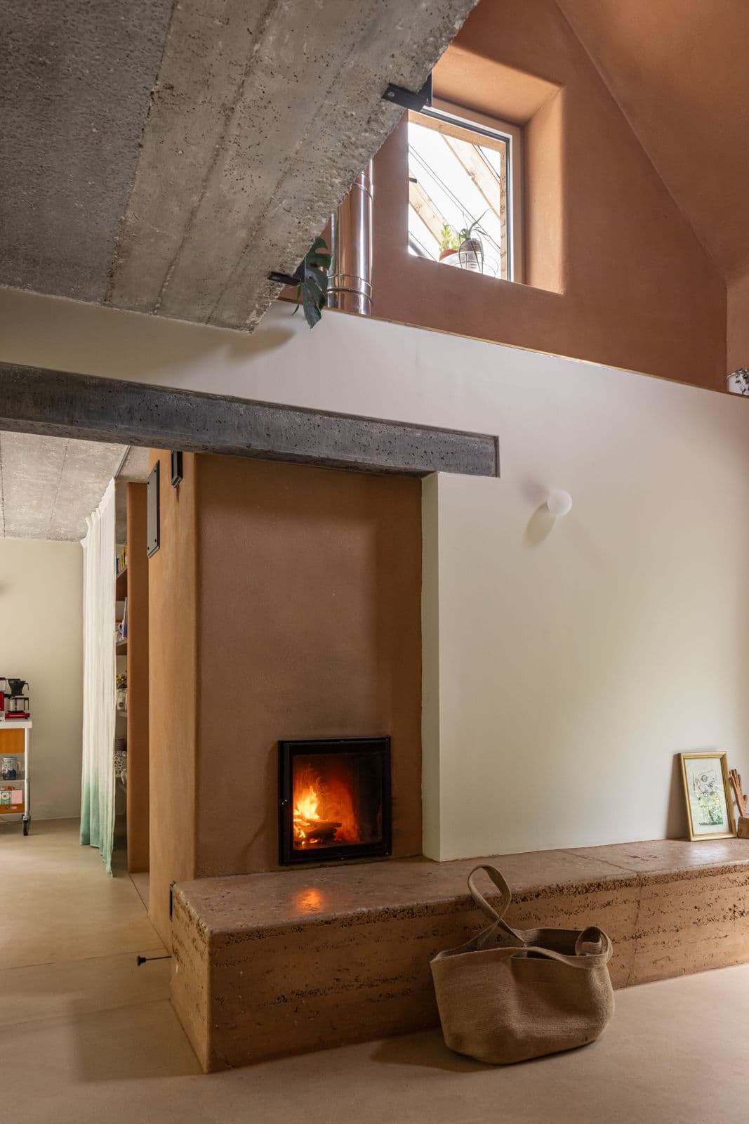 Architectural project - raw finishes, exposed concrete, indoor greenery, cozy living, living room, concrete ceiling, concrete beam, fireplace, built-in fireplace, seated hearth, homely space, subtle colors, warmth, rustic finishes, warm interior, intimate space, natural materials, casual style, natural light, inviting atmosphere, sustainable design, warm tones, eco design, neutral tones, beige, brown, concrete, light wood, compact space, simple design, rustic vibe, rustic modern, industrial touch, minimalist style, neutral palette, modern rustic, architectural details, plaster wall, energy efficient, plaster, small window, warm ambiance, terracotta wall, earthy tones, terracotta, cozy corner, textured walls, comfortable living, skylight, welcoming home