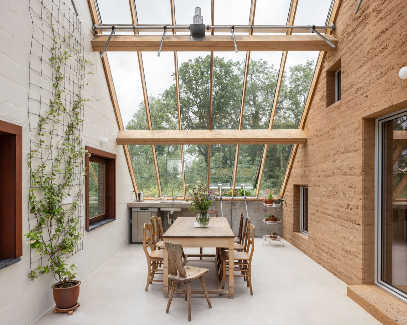 Architectural project - indoor greenery, indoor garden, wood furniture, glass roof, earth wall, kitchen area, house plants, wood, timber, natural light, sustainable design, recycled materials, glass, warm tones, neutral tones, daylight, beige, brown, concrete, farmhouse style, honey wood, wood beams, timber beams, natural textures, minimalist design, eco-friendly materials, rammed earth, modern rustic, biophilic design, eco home, sustainable home, passive house, natural ventilation, light filled, white, cozy vibe, landscape view, rustic style, open layout, open plan, dining area, inviting space, nature-inspired, bright interior, plaster walls, earthy textures, clay plaster, skylight, vertical garden