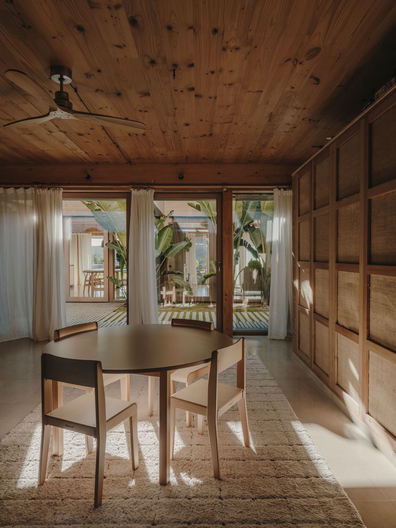 Architectural project - curtain divider, wooden chairs, cozy room, three chairs, airy room, bohemian touch, circular table, woven fabric, natural materials, sustainable materials, wood, timber, natural light, eco design, wooden ceiling, light wood, Scandinavian influence, sunlight, rustic vibe, neutral palette, minimalist design, relaxed atmosphere, cozy space, wood floor, biophilic design, large windows, beige tones, timber ceiling, warm ambiance, rustic style, open layout, open plan, living area, inviting space, earthy tones, casual dining, textured walls, wooden panels, reading nook, home office, sliding doors, natural interior, indoor outdoor flow, simple furniture, geometric table, indoor plants, woven texture