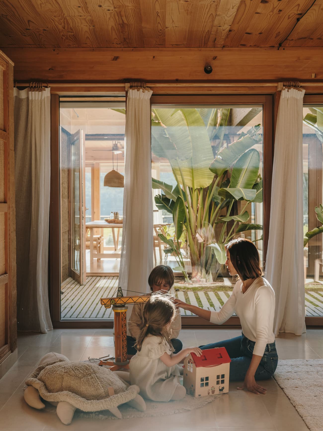 Architectural project - floor to ceiling windows, home decor, tropical plants, children playing, cozy environment, wooden toys, decoration, curtain, warm interior, wood, timber, relaxed vibe, natural light, sustainable design, glass, warm tones, neutral tones, exposed beams, daylight, beige, light wood, honey wood, Scandinavian style, biophilic design, large windows, indoor outdoor, white, Nordic design, eco friendly home, inviting home, timber ceiling, garden view, minimalist aesthetic, calm atmosphere, earthy textures, comfortable living, wood ceiling, family room, playful space, wooden furniture, sliding glass doors, play area, living space