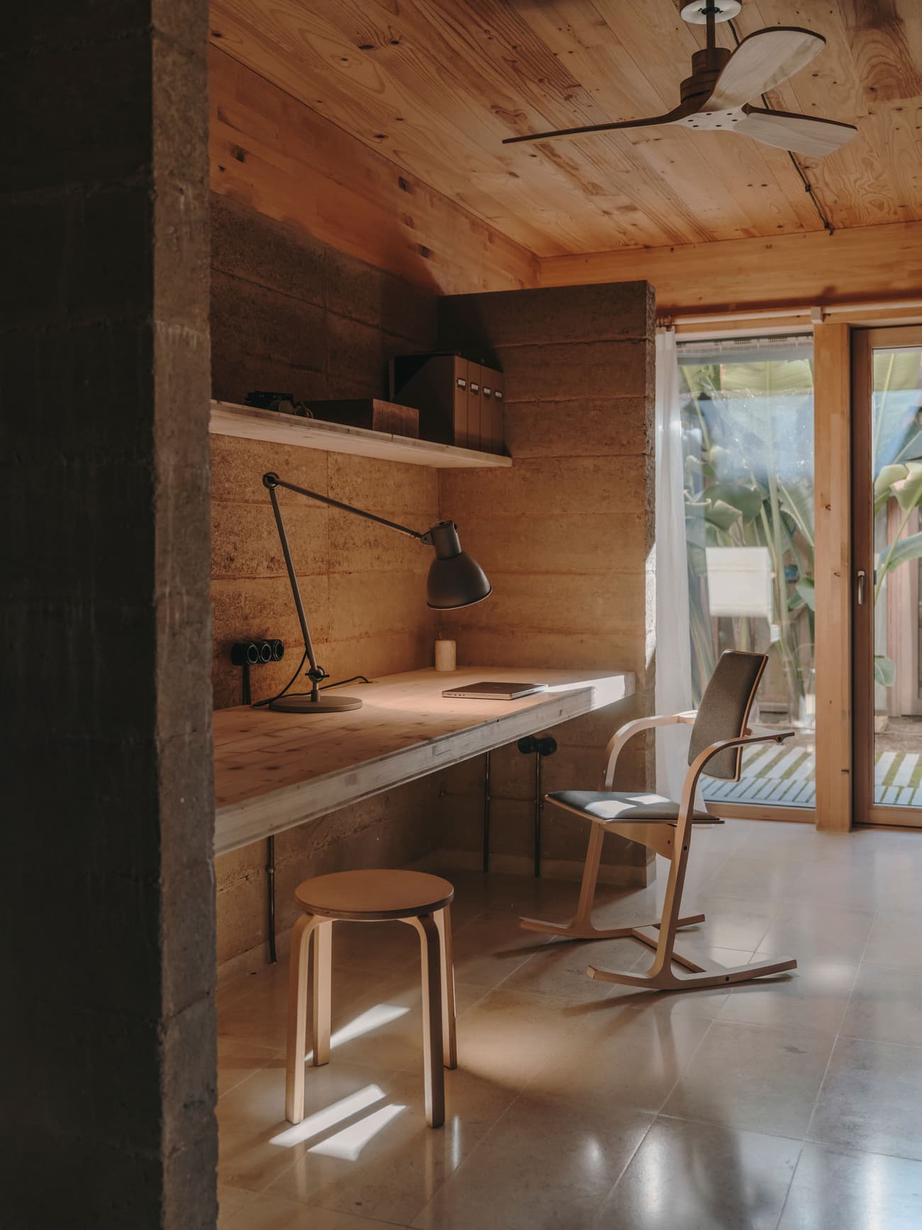 Architectural project - cozy office, warm office, wooden desk, timber desk, minimalist chair, office chair, study lamp, sliding glass door, plants, tropical view, earthy walls, calm vibe, comfortable workspace, inviting workspace, sustainable materials, wood, timber, raw materials, relaxed vibe, natural light, warm tones, natural tones, eco design, neutral tones, daylight, beige, concrete, light wood, functional design, compact space, simple design, rustic modern, serene atmosphere, Scandinavian style, minimalist style, workspace, rammed earth, modern rustic, passive house, natural ventilation, indoor outdoor, Nordic design, timber ceiling, garden view, textured walls, home office, wood ceiling, interior design, Japandi, organic textures, wooden stool, home renovation, reading lamp