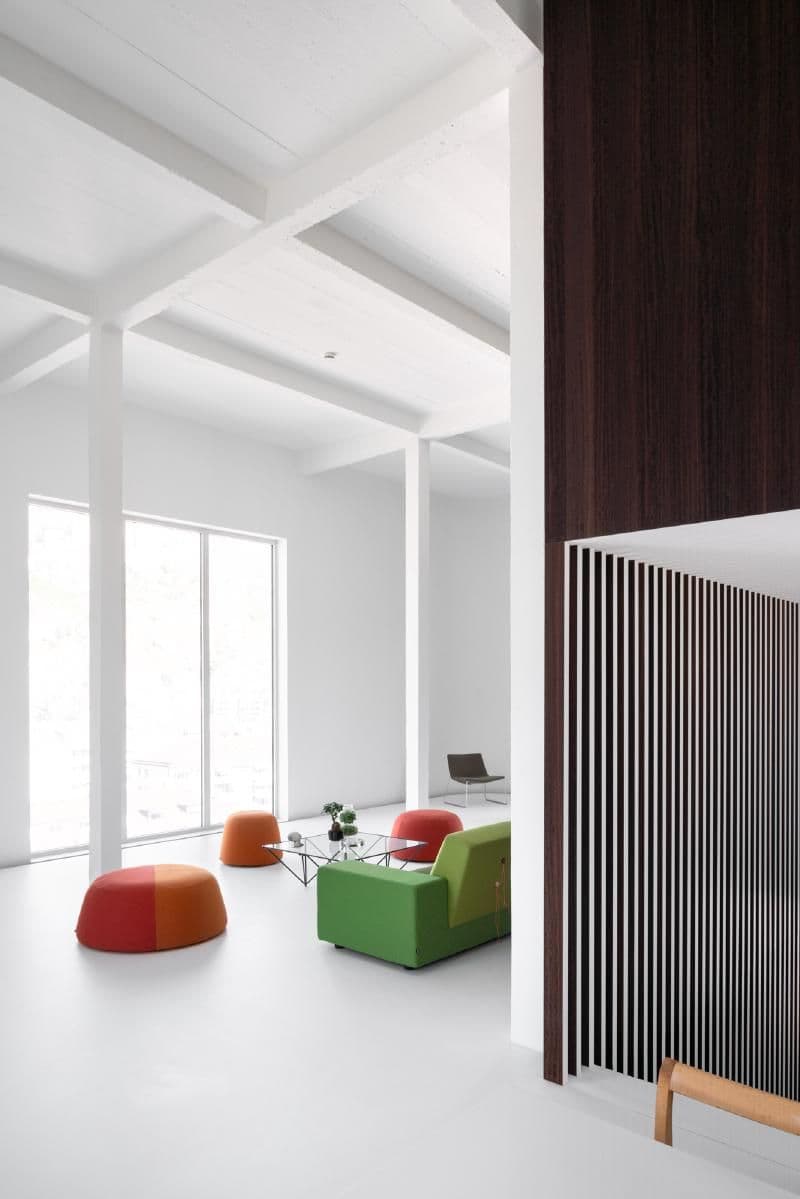 Architectural project - lounging area, modern decor, colorful furniture, vibrant seating, simplicity, green sofa, orange pouf, red pouf, striped accents, eco-friendly design, wood, timber, natural light, green, neutral tones, ceiling beams, daylight, gathering space, spacious layout, brown, modern aesthetic, functional design, casual seating, social space, relaxed atmosphere, orange, clean lines, white walls, white, Nordic style, large window, cozy corner, modern living room, Scandinavian design, bright interior, sleek design, home design, open space, contemporary vibe, airy feel, vertical slats, wood paneling, minimalist interior, residential interior, red