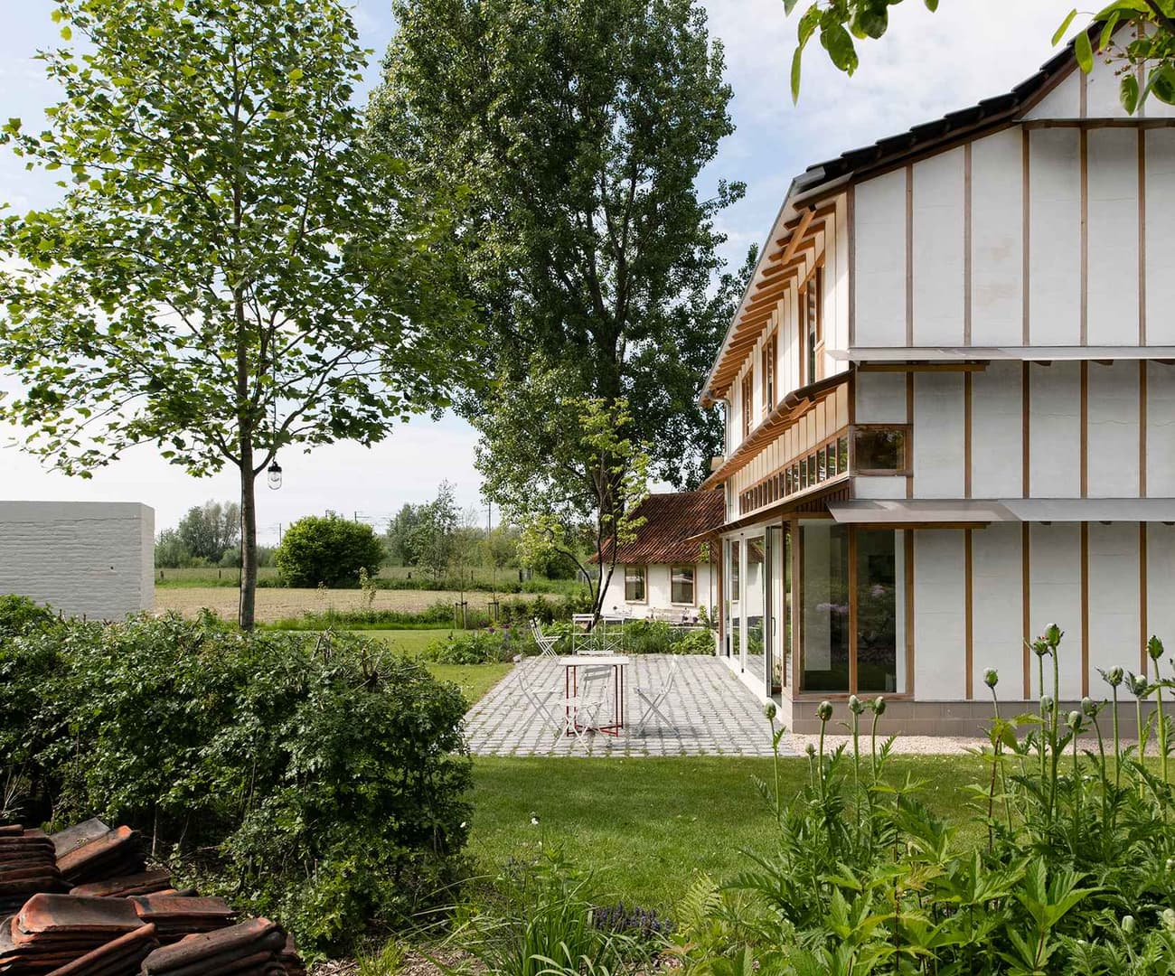Architectural project - wood, timber, outdoor space, outdoor living, sustainable design, glass, natural tones, green, light wood, farmhouse style, rustic charm, outdoor seating, low carbon, biophilic design, passive house, white facade, large windows, wood cladding, white, cozy vibe, inviting home, timber house, rural home, earthy tones, garden retreat, rural setting, landscape design, lush garden, natural environment, garden view, nature integration, eco house, serene landscape, residential home, countryside house, timber framing, patio area, comfortable living, traditional house, paved patio, grassy yard, pastoral scene, wood planks, country living, green surroundings