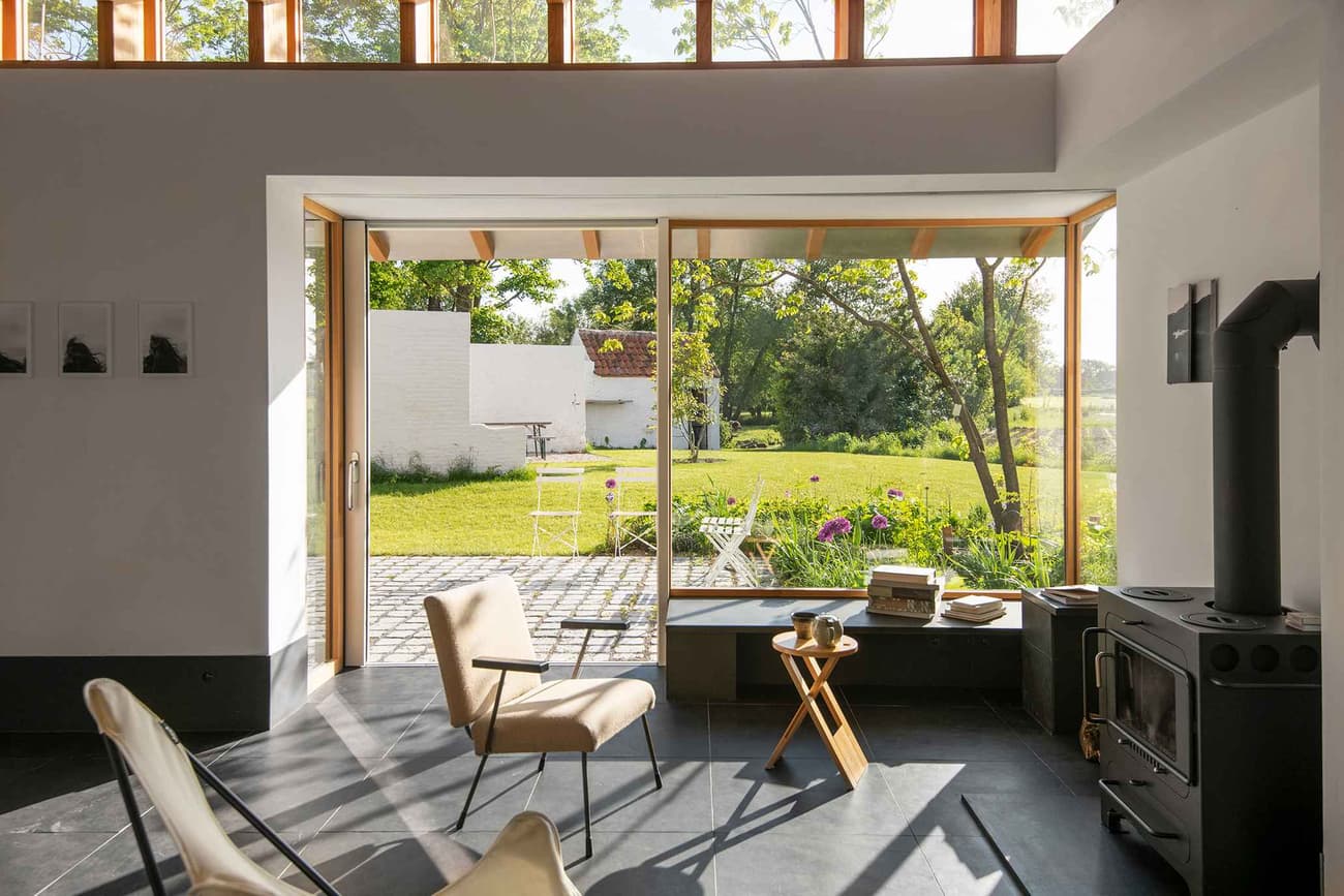 Architectural project - minimal design, cozy interior, eco-friendly design, natural light, inviting atmosphere, passive design, eco-conscious, daylight, multifunctional space, Scandinavian style, neutral palette, light tones, modern rustic, sustainable home, large windows, indoor outdoor, landscape view, wood accents, nature-inspired, relaxing space, outdoor connection, rural setting, garden view, nature immersion, comfortable seating, minimalist aesthetic, bright interior, glass door, open living, lounge area, wooden frames, timber frames, Nordic vibes, warm living, sunlit room, fluid transition, modern cottage, simple decor, black floor, slate floor, garden seating, compact living, efficient layout, small table, modern stove, hearth, indoor warmth