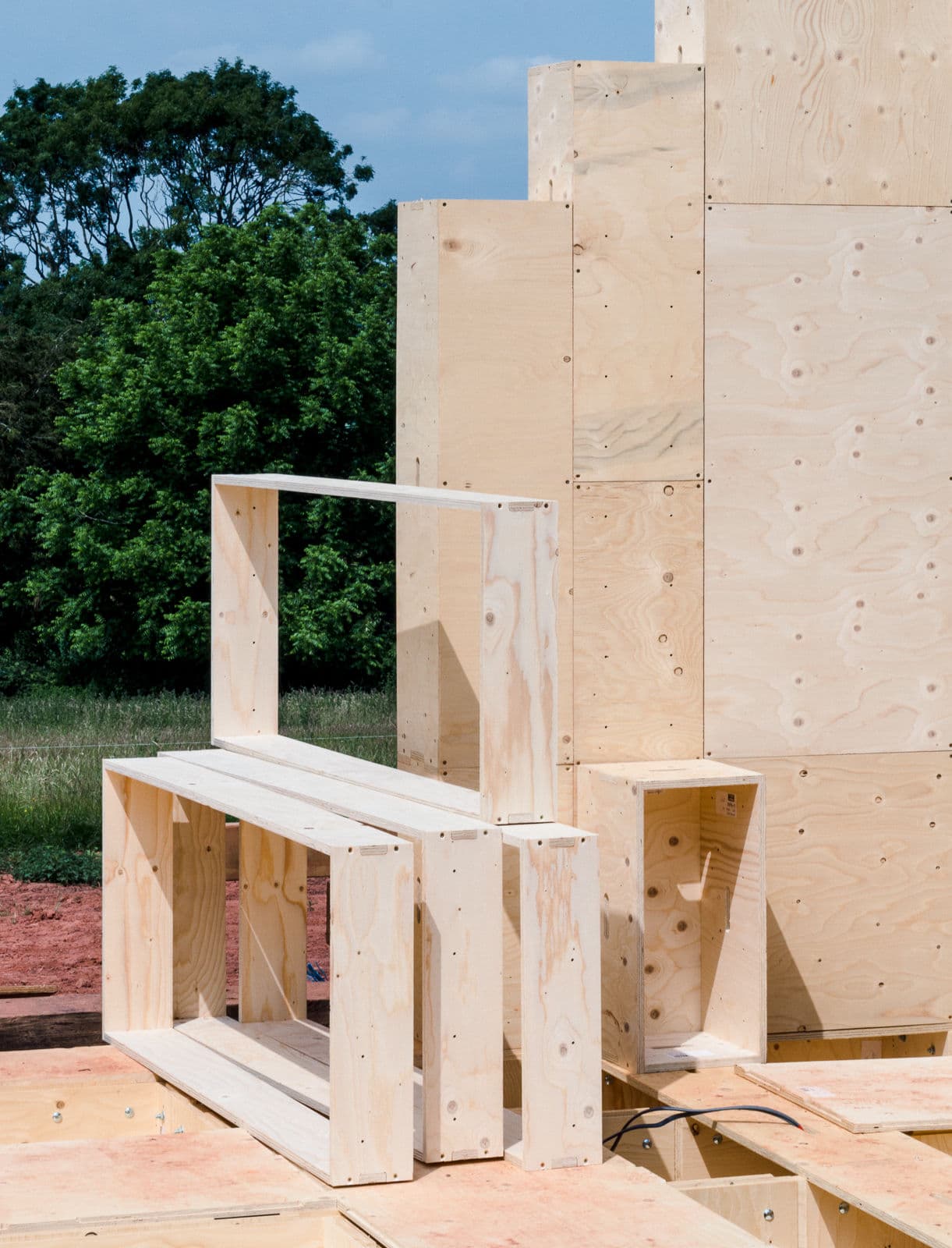 Architectural project - field setting, simple geometry, built environment, plywood structure, wood design, raw plywood, wood assembly, structural plywood, stacking design, creative structure, landscape interaction, yellow wood, assembly art, natural materials, sustainable materials, timber, functional art, outdoor space, natural tones, modular design, eco design, innovative design, architectural installation, outdoor installation, modern construction, plywood, beige, light wood, engineered wood, eco friendly, natural setting, timber construction, neutral palette, minimalist design, construction detail, prefabricated elements, low carbon, environmental design, landscape design, greenery, wooden panels, open space, texture play
