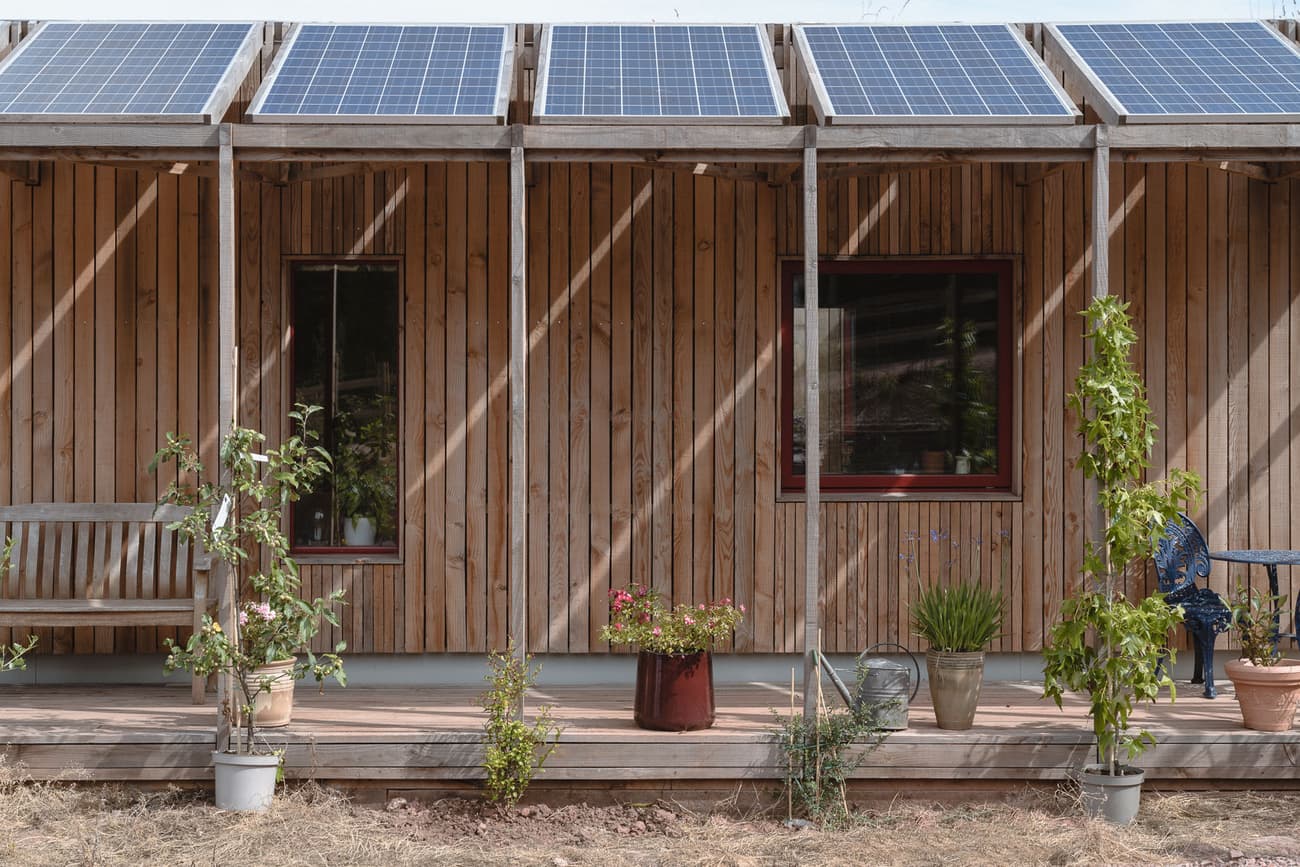 Architectural project - minimalist home, solar panels, solar energy, countryside style, country house, patio seating, wooden planks, veranda, plant pots, outdoor plants, energy conscious, cozy facade, perimeter garden, natural materials, wooden bench, wood, timber, steel, relaxed vibe, sustainable design, glass, green, neutral tones, timber structure, beige, brown, wood facade, low carbon design, eco friendly, wood exterior, natural setting, simple design, natural textures, eco home, passive house, natural ventilation, timber cladding, inviting home, timber house, energy efficient, rustic style, earthy tones, warm atmosphere, rustic facade, casual outdoor, rural setting, exterior view, green design, rustic home, wooden structure, vertical cladding, sustainable living, garden space