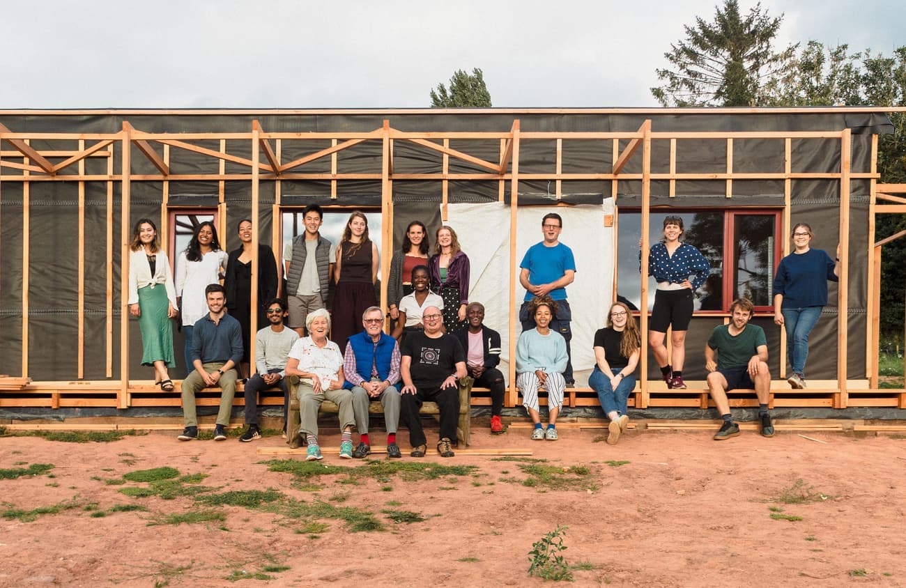 Architectural project - off-grid, DIY construction, biophilic touch, group photo, collaborative build, group activity, greenery backdrop, innovation in architecture, social project, natural materials, wood, timber, passive design, sustainable design, modular design, beige, brown, architectural framework, eco friendly, modern design, natural setting, timber construction, natural textures, minimalist structure, prefab elements, eco build, low carbon, circular design, earth tones, clean lines, timber cladding, simple facade, energy efficient, outdoor gathering, rural setting, exterior view, open air, wooden facade, community project, versatile space