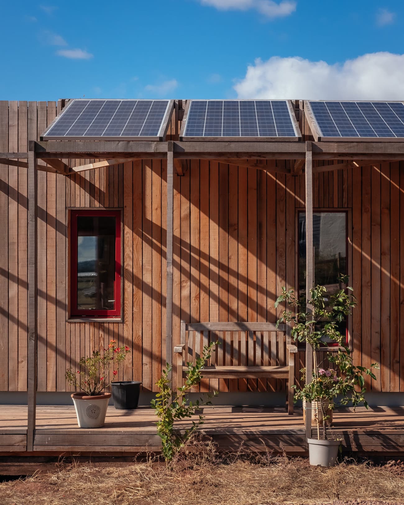 Architectural project - natural shade, warmth, solar panels, potted plants, contemporary barn, serene ambiance, plant pots, porch, sunny exterior, exterior sitting area, minimal design, natural materials, wooden bench, wood, timber, natural light, shadow play, sustainable design, warm tones, eco design, simple structure, brown, wood facade, timber facade, daylight optimization, outdoor leisure, natural textures, outdoor seating, raw wood, green architecture, modern rustic, eco home, cozy vibe, countryside home, energy efficient, rustic style, earthy tones, rural setting, inviting exterior, wooden deck, passive solar, vertical cladding, sustainable living